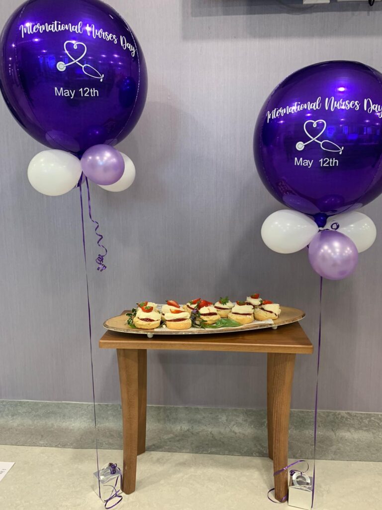 Colour photograph of a platter of scones with cream, jam and strawberries and two large purple balloons with the text 'International Nurses Day May 12th' printed on them