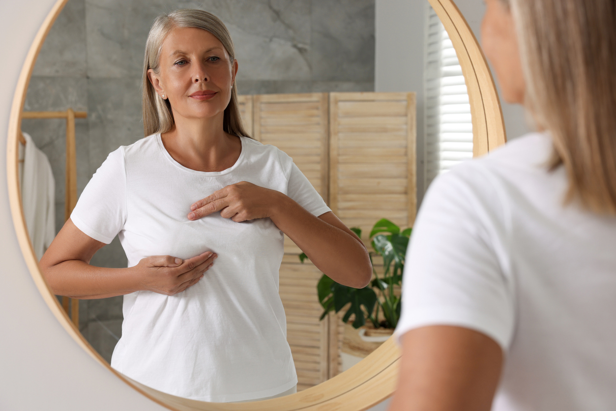 Beautiful senior woman doing breast self-examination near mirror in bathroom
