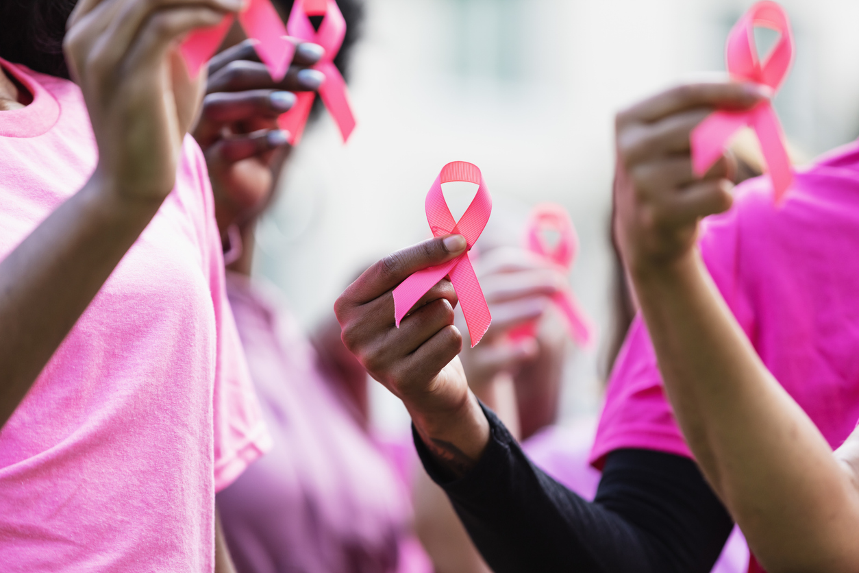 A pink breast cancer awareness ribbon held aloft in the fingers of a mid adult African-American woman. The focus is on her hand and the ribbon. She is surrounded by other women holding ribbons.